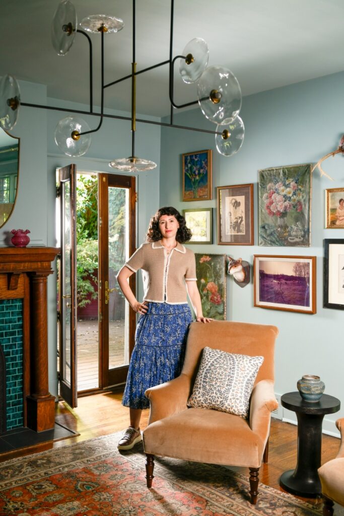 Interior designer Michelle Gage standing in a light blue living room featuring a gallery wall of fine art, an orange velvet armchair, and a modern glass chandelier.