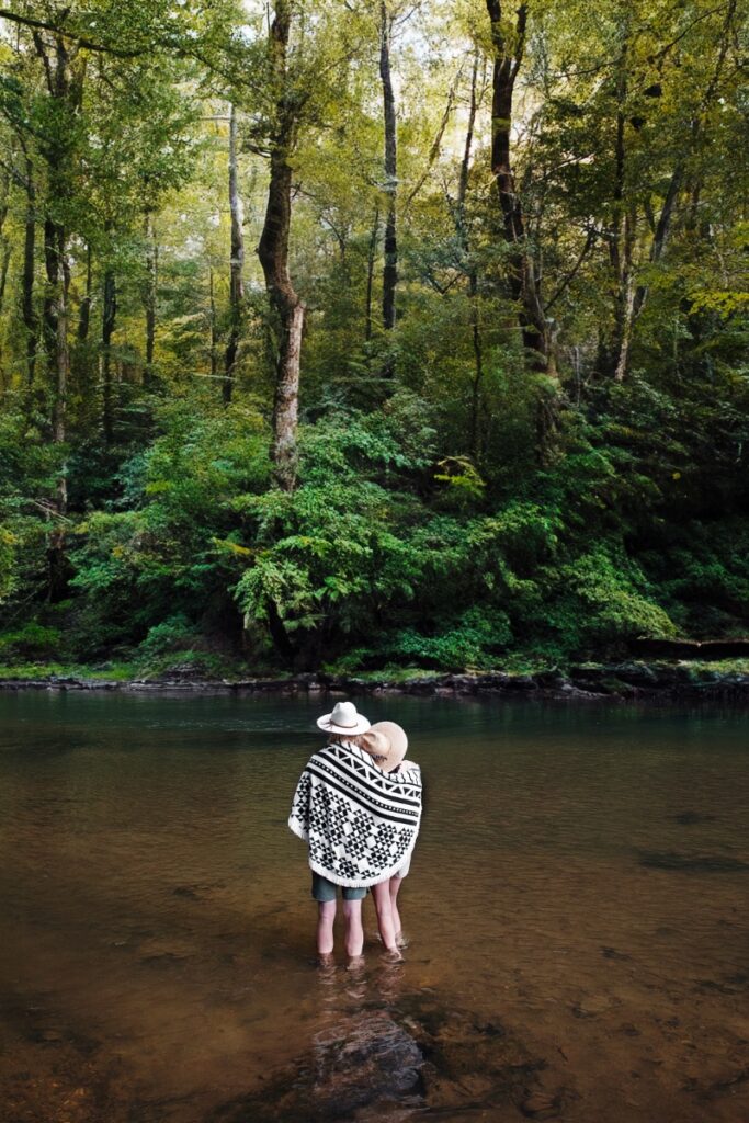 A couple wrapped in a black-and-white patterned blanket standing in a shallow river surrounded by lush green forest during a romantic trip to the Georgia highlands.