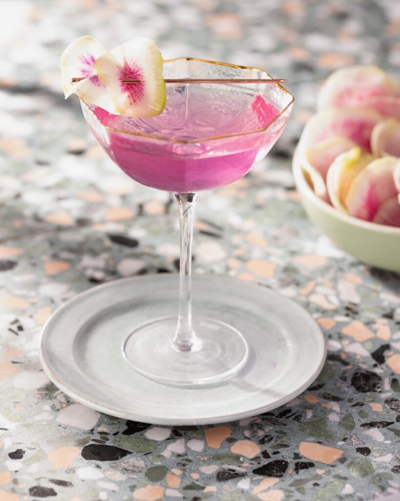 A tall pink martini cocktail with a watermelon radish on a tooth pick as a garnish.