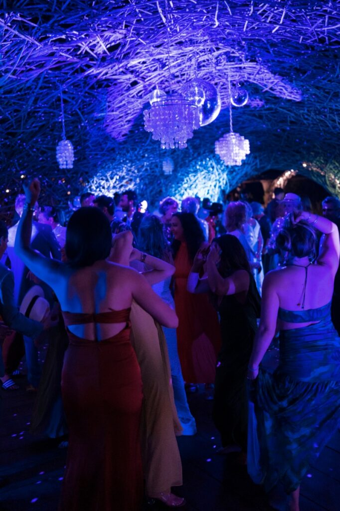 Wedding guests dancing under vibrant blue and purple lights in a rustic, woven tunnel structure during an evening reception in Todos Santos, Mexico.
