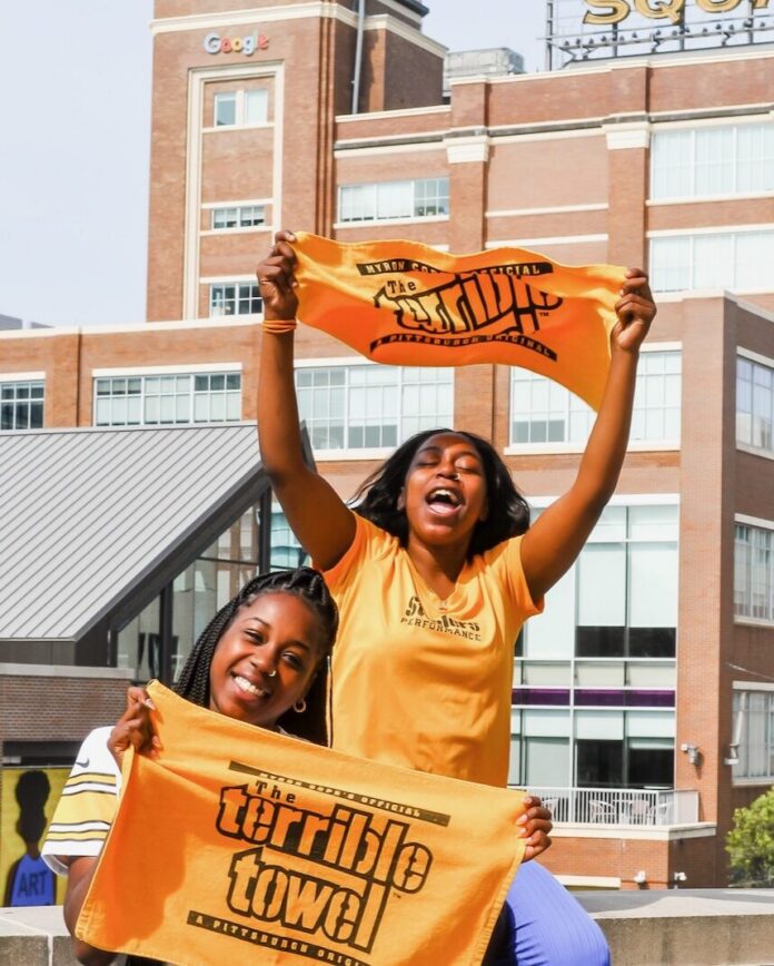Two women in Steelers shirts hold up Terrible Towels in front of Bakery Square in Pittsburgh.