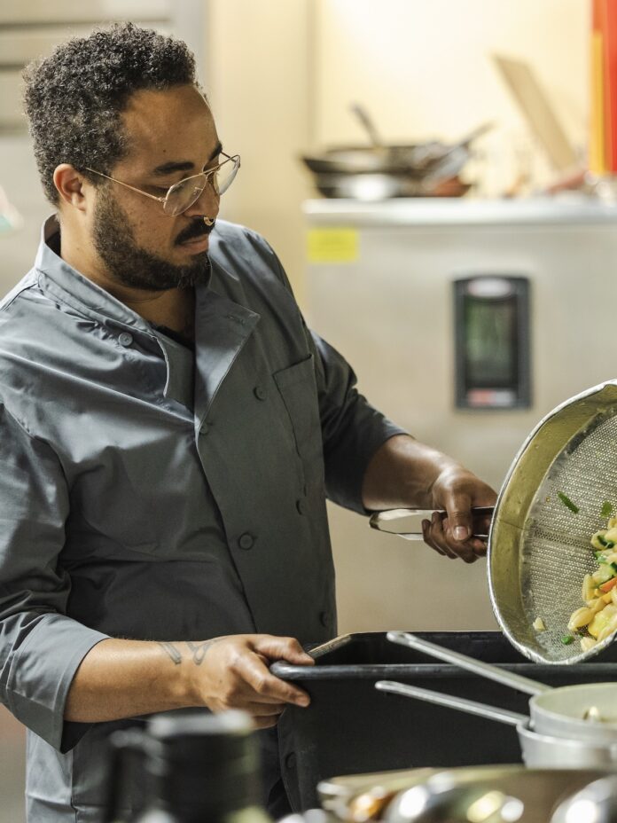 A man in a blue chef coat dumping pasta from a strainer into a pot.