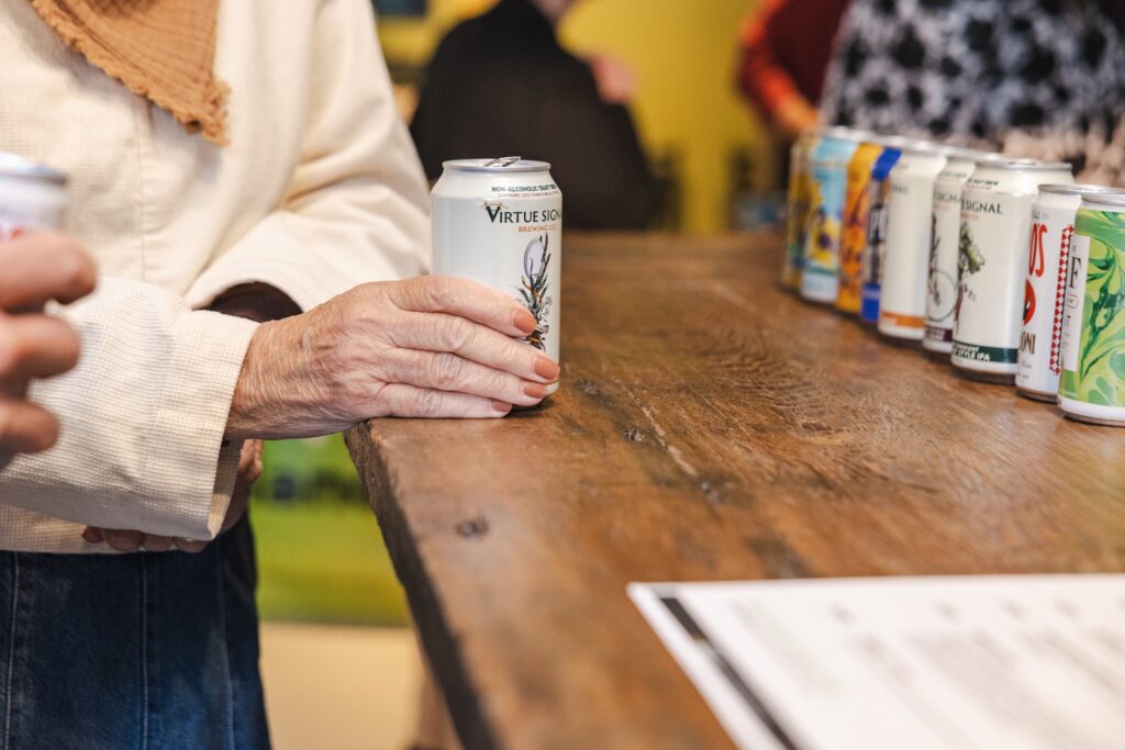 An older woman holding a can of n/a Virtue Signal Brewing Co. on a wooden bar with cans in the background.