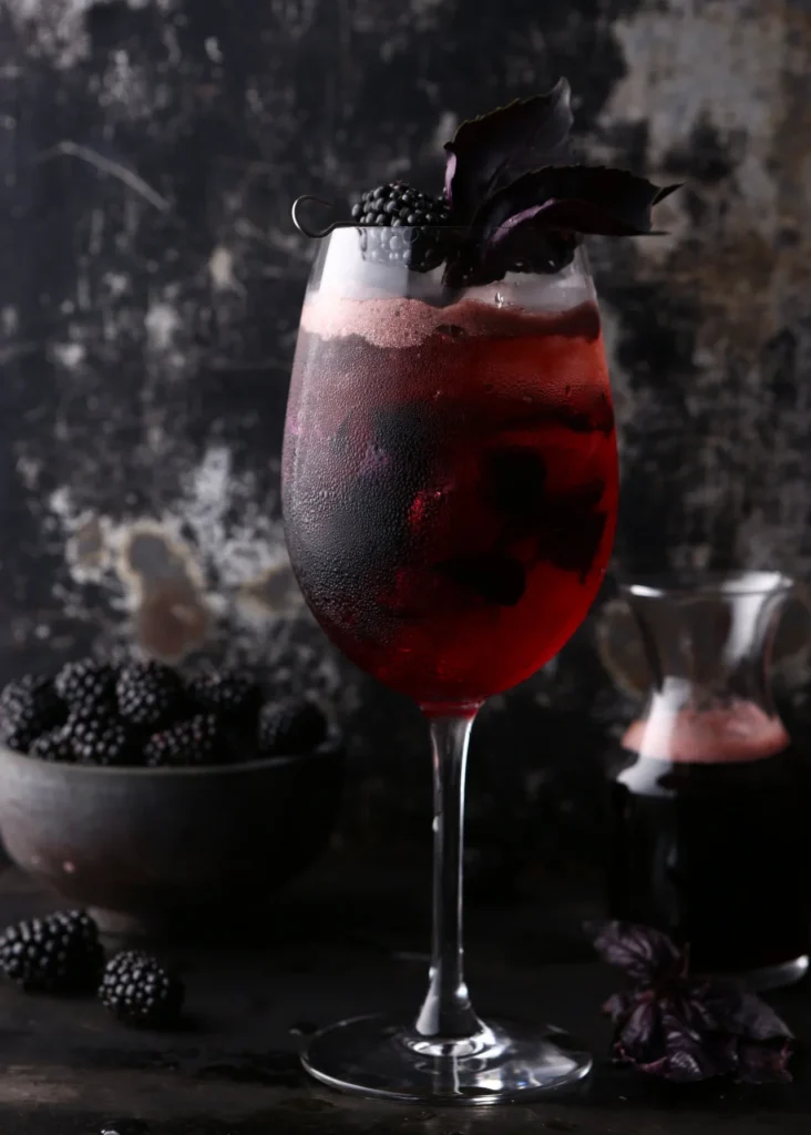 A wine glass holds a Blackberry Crush Spritz with blackberries in the liquid and as a garnish all against a dark background.
