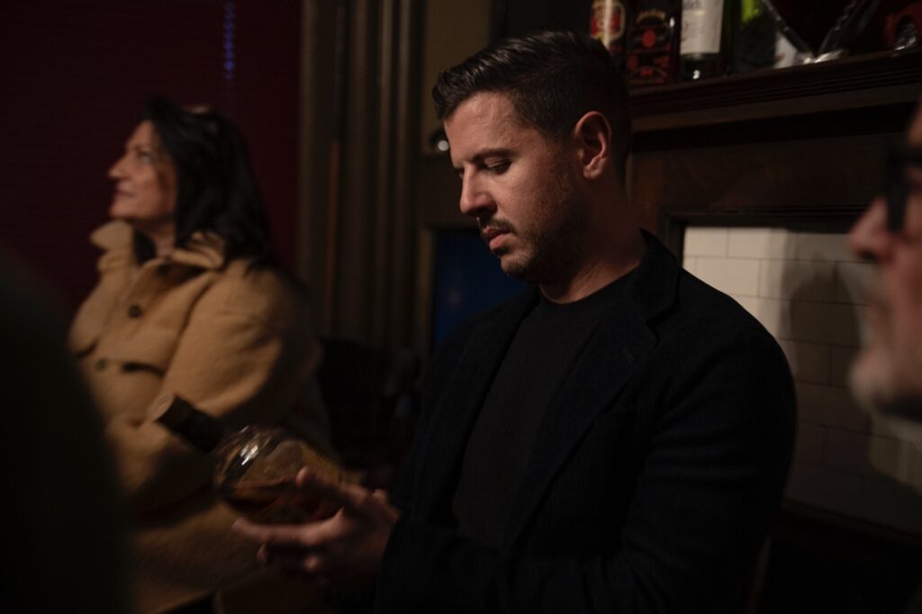 A man in a black shirt looks at a bottle of scotch in his hands.
