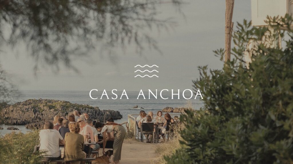 The Casa Anchoa logo and type on top of a photo of people enjoying dinner on the coast. 