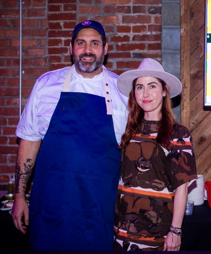 Chef Fernando Canales in his blue apron and Meredith Boyle from Balvanera stand together in front of a brick wall.