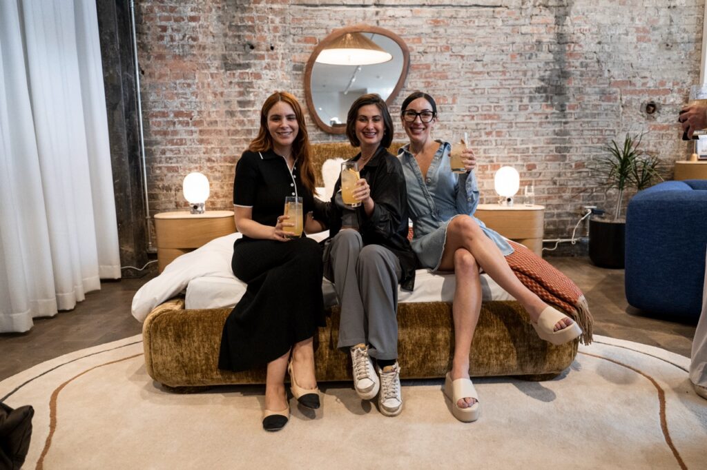 Two woman in button up dresses sit on either side of a woman in a leather jacket on a bed.