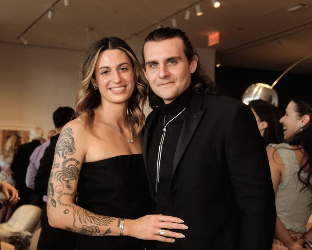 A woman with tattoos on her arm in a black dress stands with a man in a dark suit jacket and bolo tie.