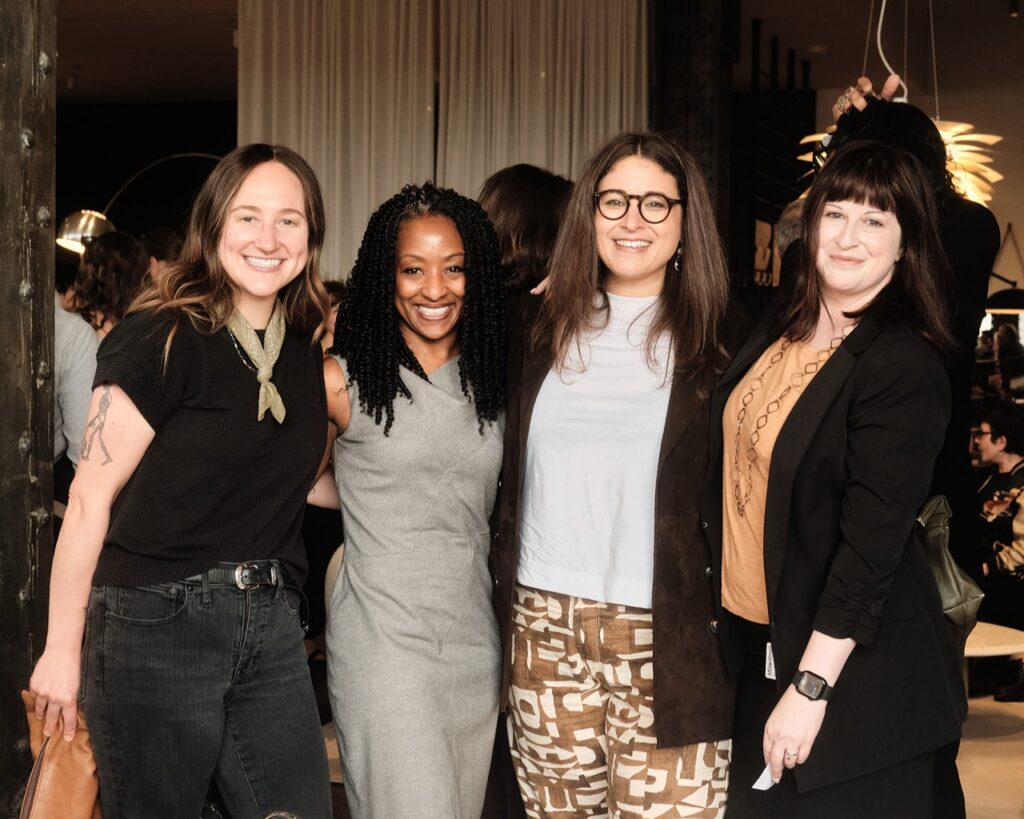 Four women in dress clothes smile with their arms around each other.