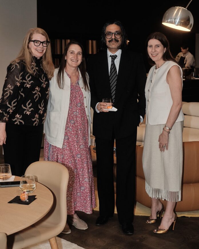Three women and a man in a suit and tie stand in front of a couch.