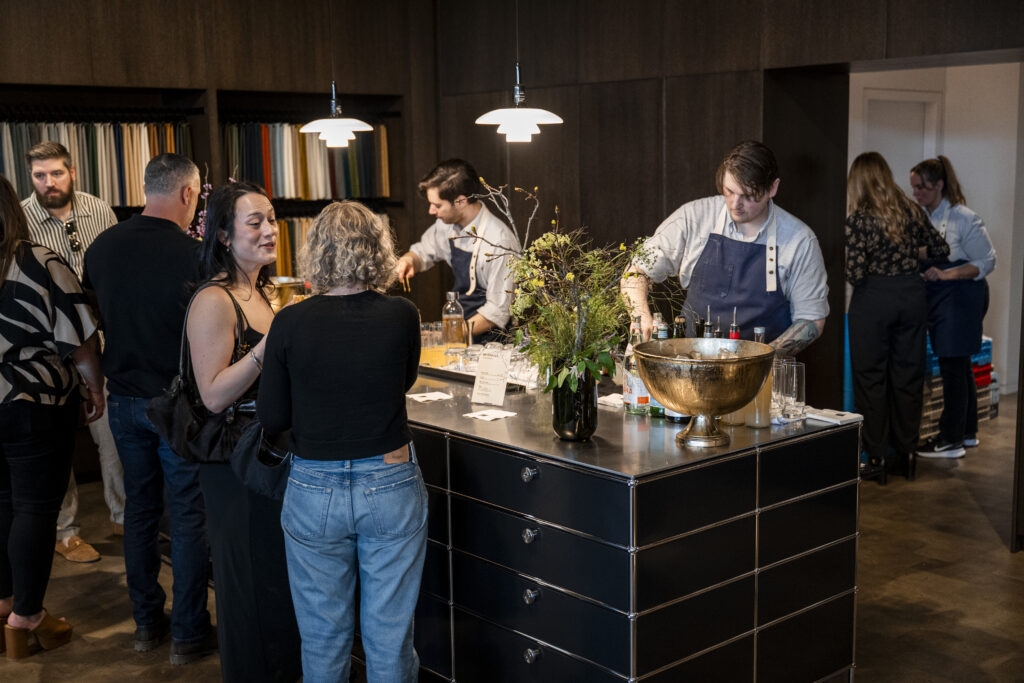 People gather around a counter where two men in aprons are serving drinks.