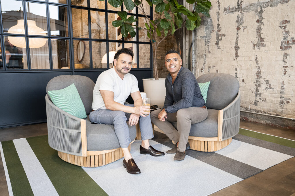 A man in a white shirt and a man in a blue shirt sit on grey chairs with a plant behind them.
