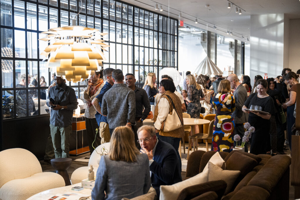 A crowd of people talk to one another with the showroom windows in the background and furniture throughout.