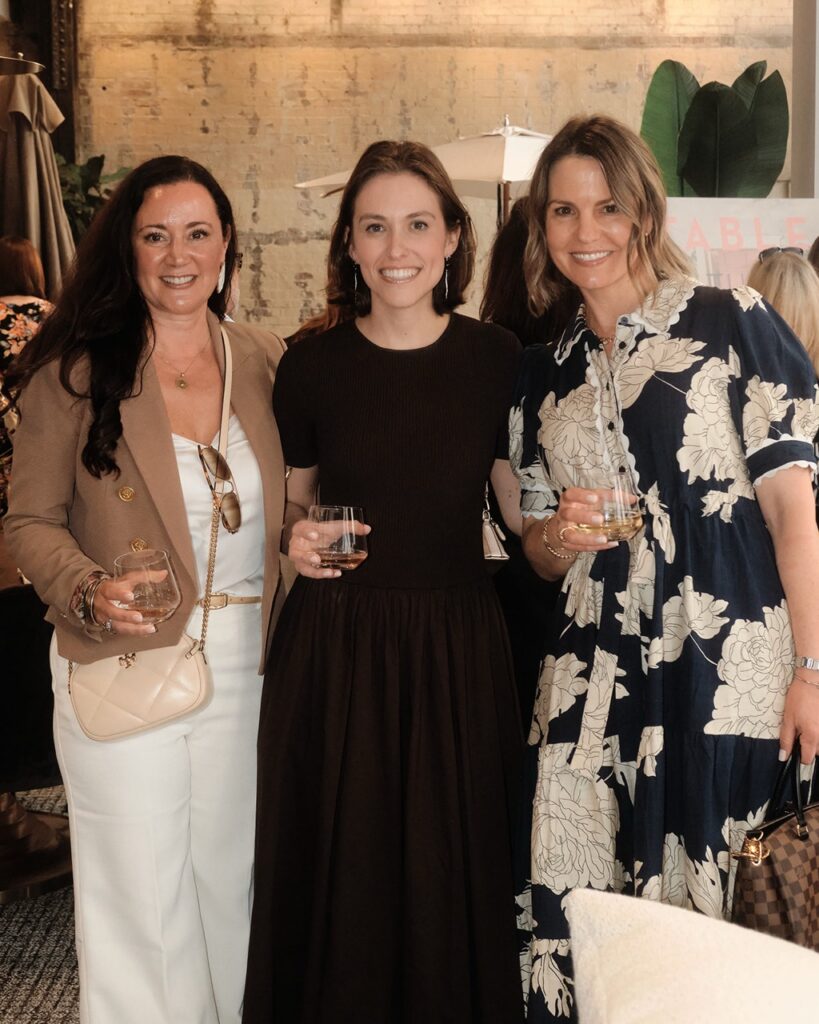 A woman in a cream jacket and white jumpsuit stands with a woman in a long black dress and a woman in a long navy dress with white flowers.