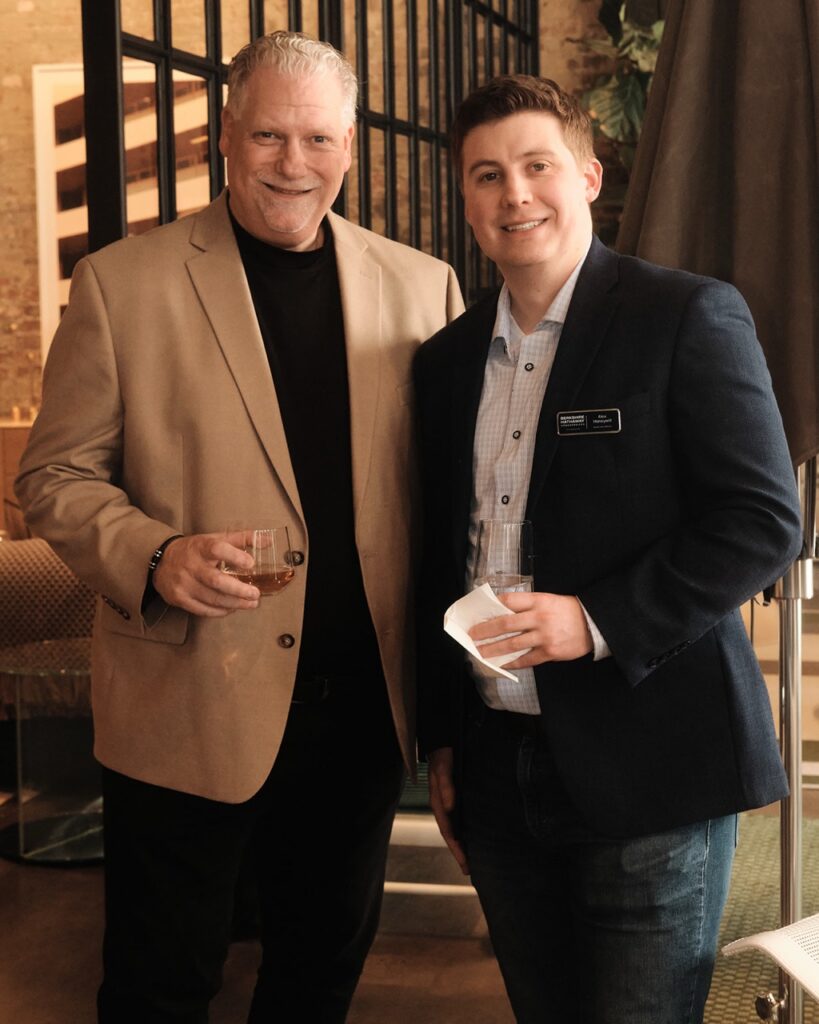 A man in a neutral tone suit jacket stands beside a man in a dark blue suit jacket with drinks in hand.
