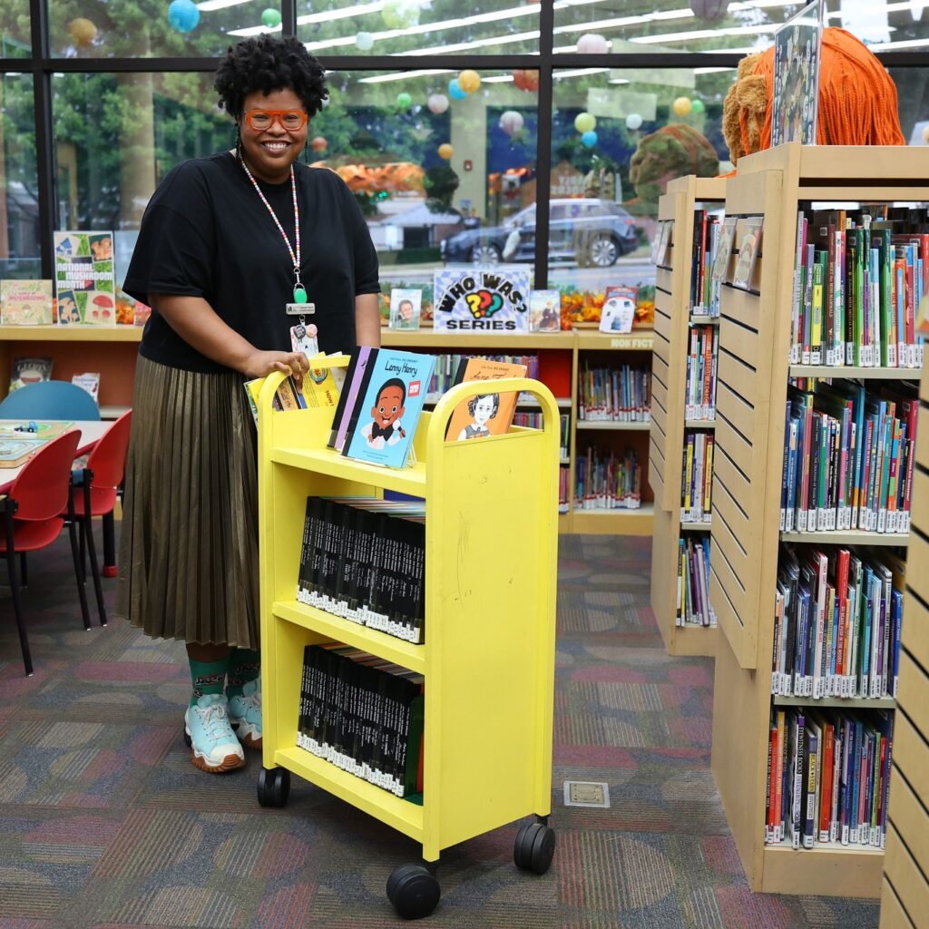 A librarian pushes a yellow cart in the library with children's books on it.