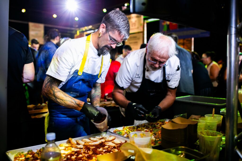 Two men in aprons dress samples of food on a table.