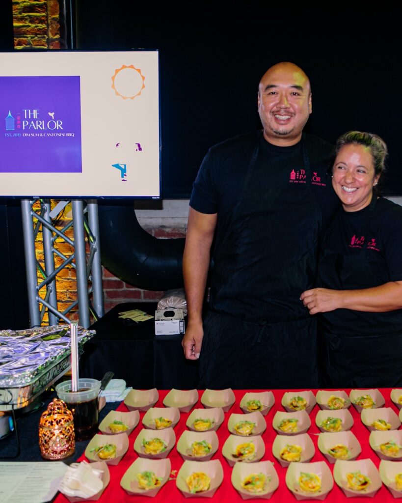 A man and woman stand arm in arm behind a table of samples.