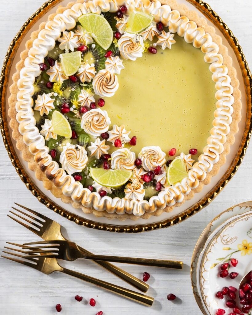 A kiwi and lime tart in a pie tie with meringue, lime wedges, kiwi, and pomegranate seeds on top.