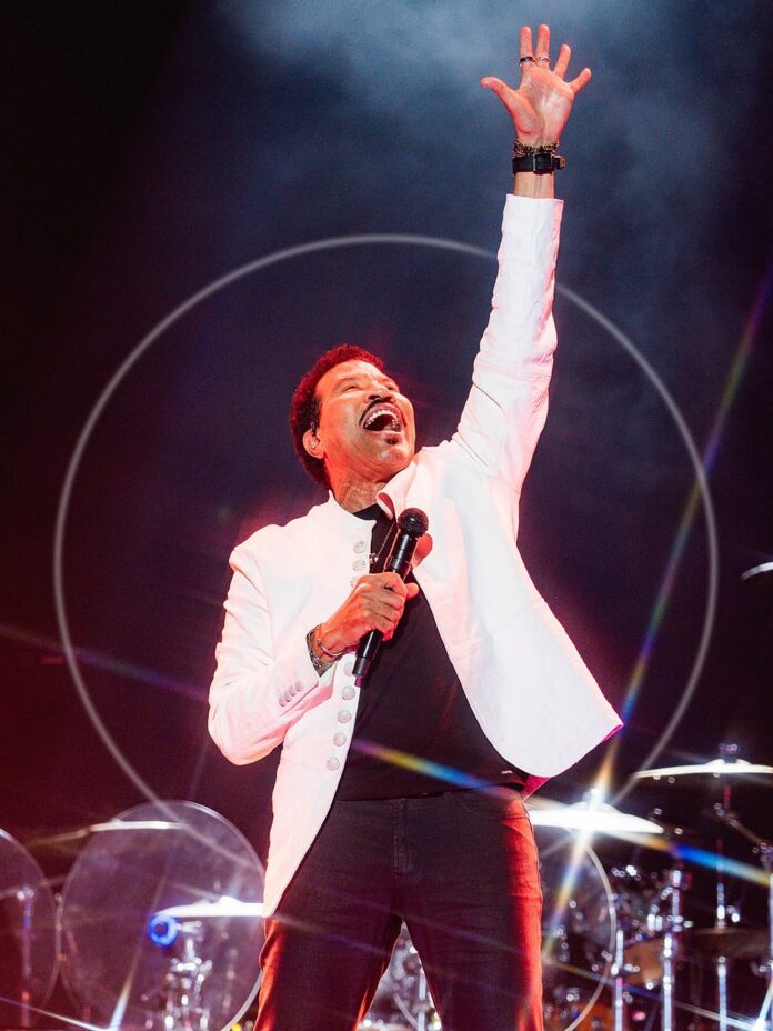 Lionel Richie in a white jacket singing on stage at a concert with his hand reaching up to the sky.