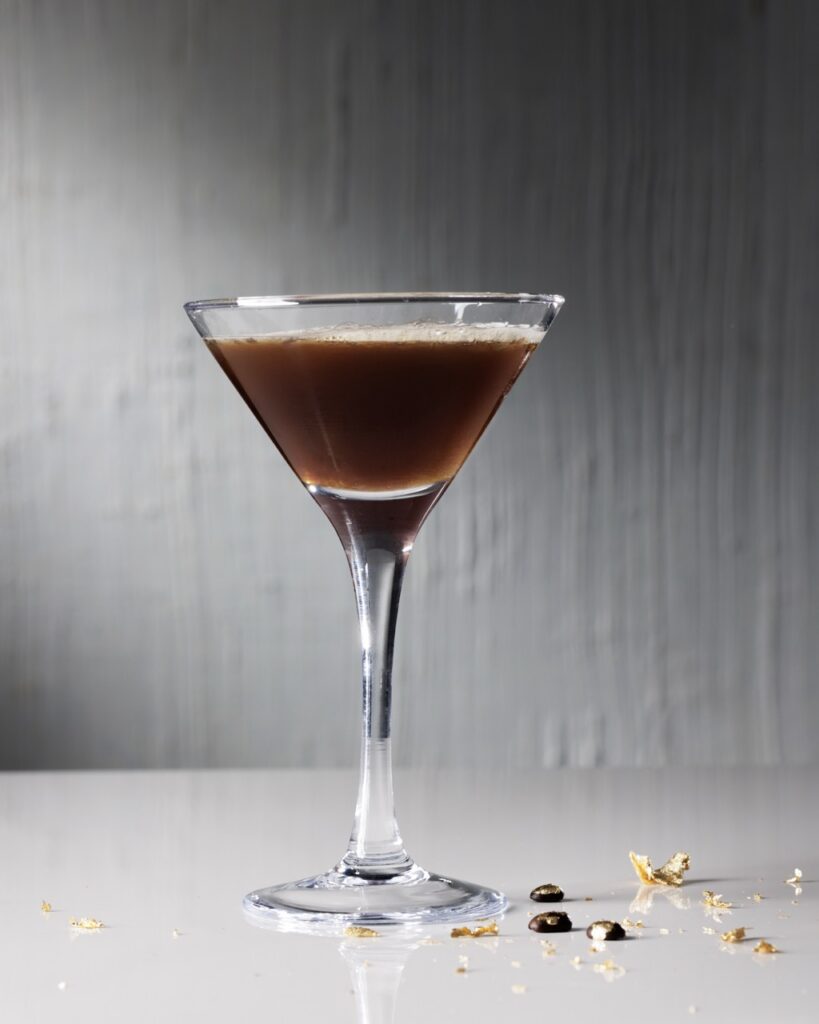 Miranda's espresso martini in a tall glass with gold leaf coffee beans on the white table below it.
