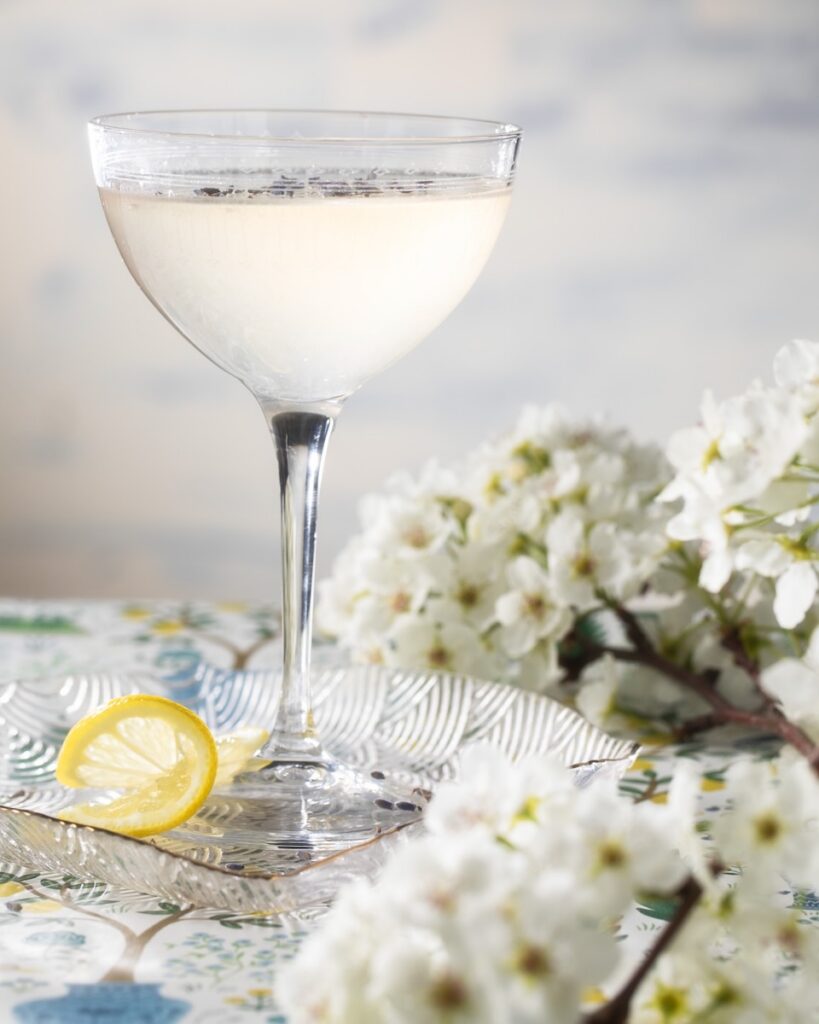 In a coupe glass on a table cloth sits a Lavender Lemon Drop for Mother's Day as white flowers and a lemon twist lay beside the glass.