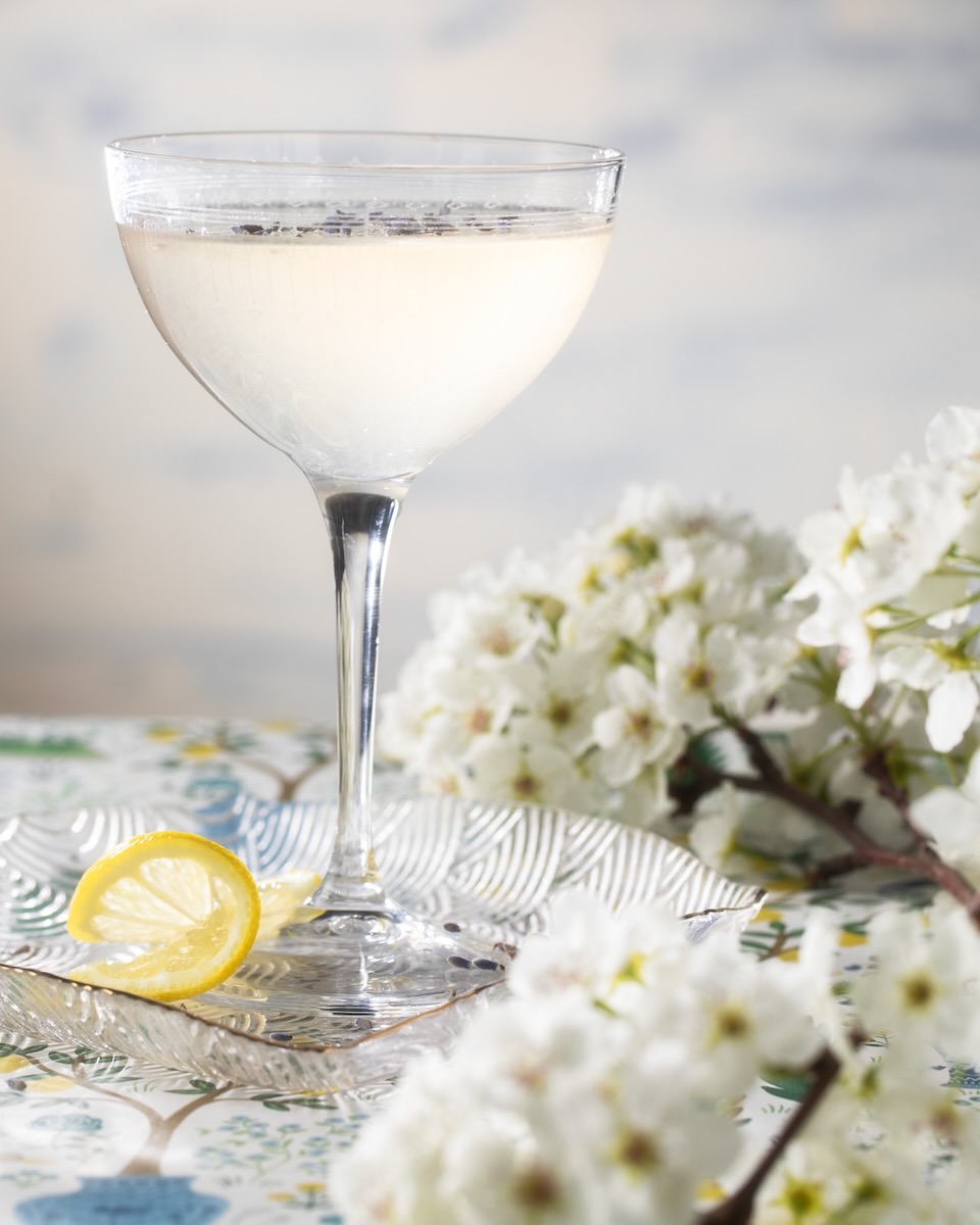 In a coupe glass on a table cloth sits a Lavender Lemon Drop for Mother's Day as white flowers and a lemon twist lay beside the glass.