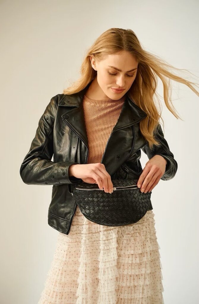 A woman in a black leather jacket unzips a black leather purse at her waist. 