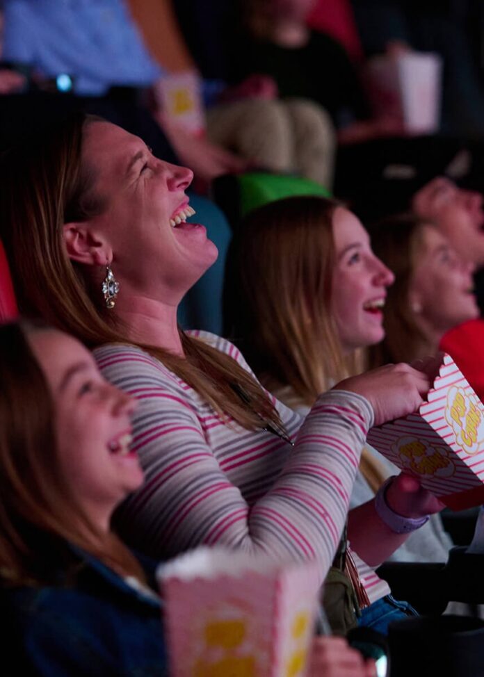 A woman at the movies laughs as she watches the screen and eats popcorn.