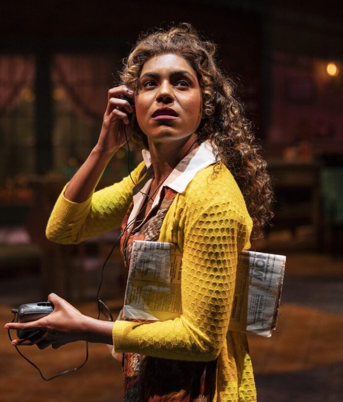 A woman in a yellow sweater holds a radio up to her ear, a newspaper under her arm.