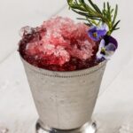 A pomegranate mint julep in a silver tin julep cup with crushed ice and a sprig of rosemary.
