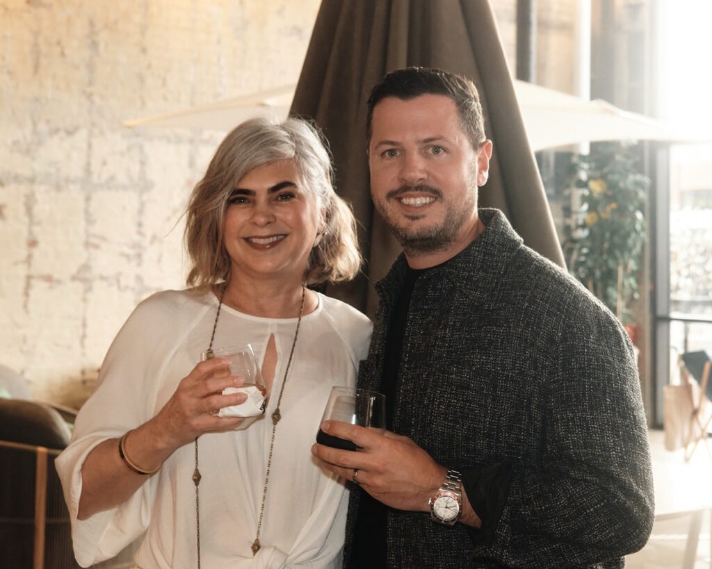 A woman in a white shirt and a man in a tweed jacket smile with drinks in hand.