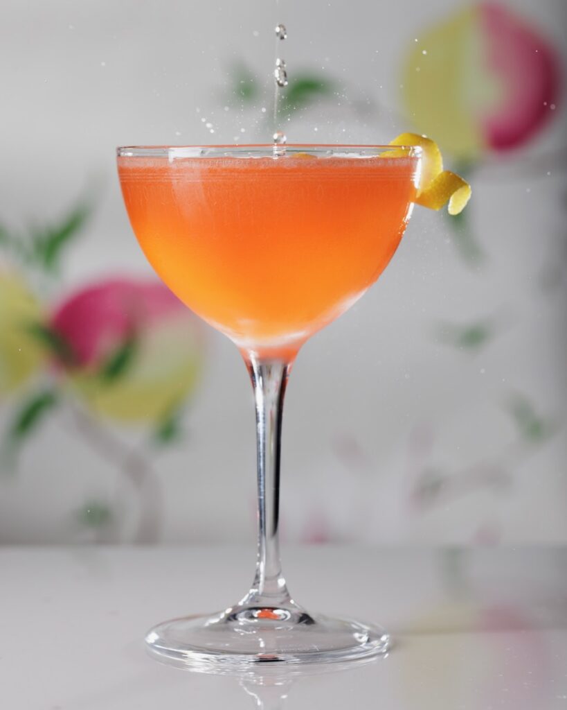 An orange color cocktail in a tall glass with a lemon twist garnish and a floral backdrop behind it.