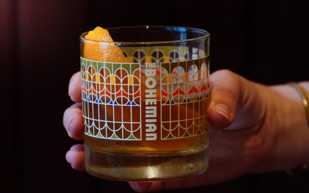 A person holds a short cocktail glass with The Bohemian logo on the outside of the glass.