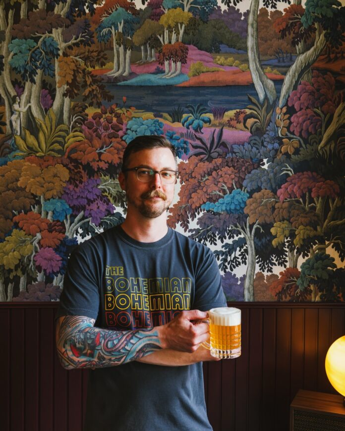 The Bohemian Bar Northside TABLE Magazine A man in a Bohemian shirt holds a pint of beer in his hand standing in front of a painted wall.