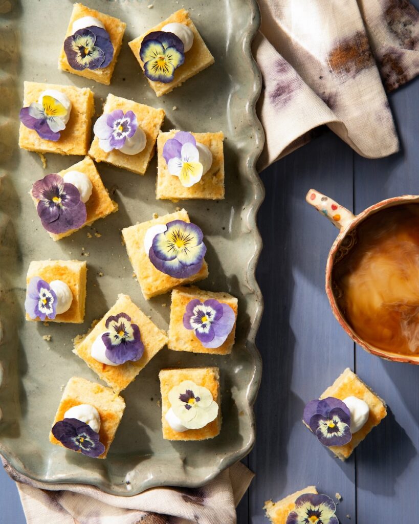 Small yellow triple citrus shortbread squares link a parchment paper baking sheet with dollops of whipped cream and purple edible flowers on top.