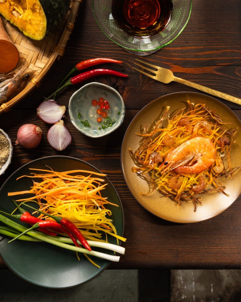 julienned vegetables sit on a green plate beside other ingredients and a brown plate with a large Filipino fritter with shrimp on top of it.
