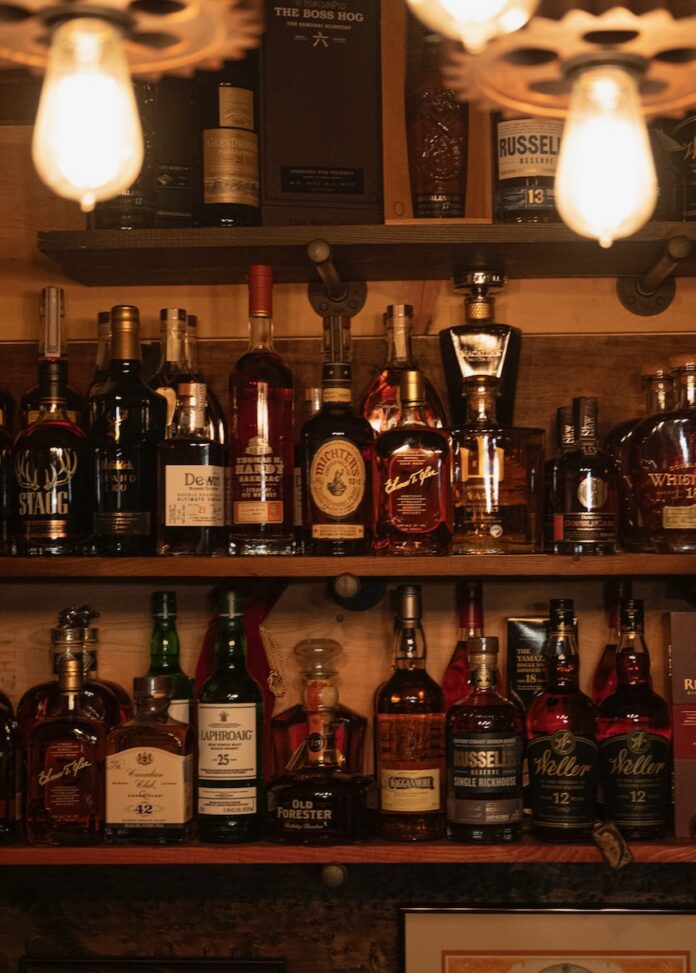 Why Scotch Tastes Different to Everyone (And How You Can Learn to Enjoy It) Two selves full of bottles of Scotch sit below two antique light bulbs behind a bar.