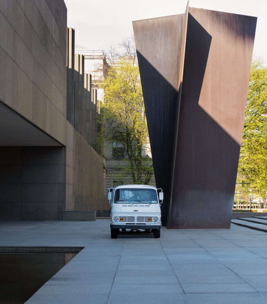 A white truck beside a large sculpture of two metal panes outside.