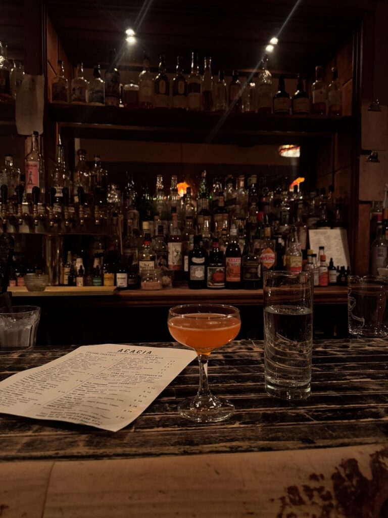 A dark bar with plenty of bottles behind it and a pink drink sitting on the wood bar. 