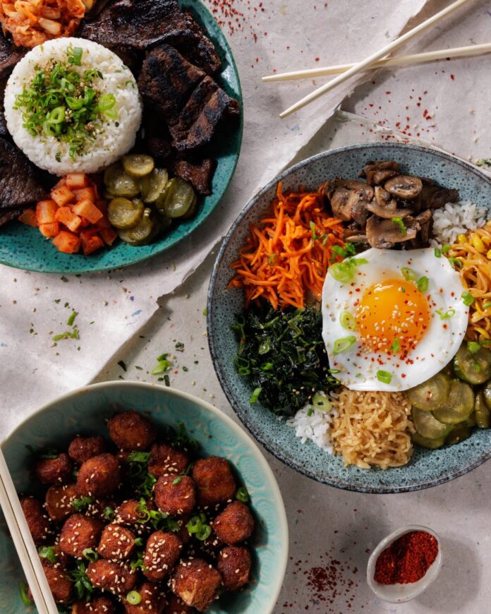 A white table with three blue bowls on it with Asian bulgogi, poke bowl with egg on top, and small fried balls with chopsticks on the table.