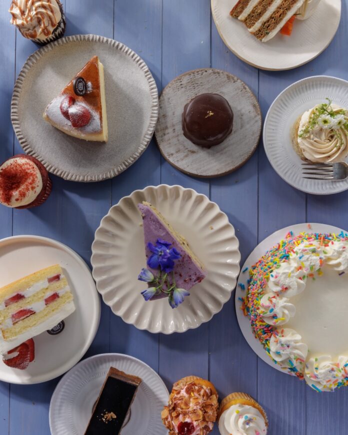 Plates full of different sices of cake like strawberry, carrot, matcha, confetti sprinkles, and other types on a purple picnic table.