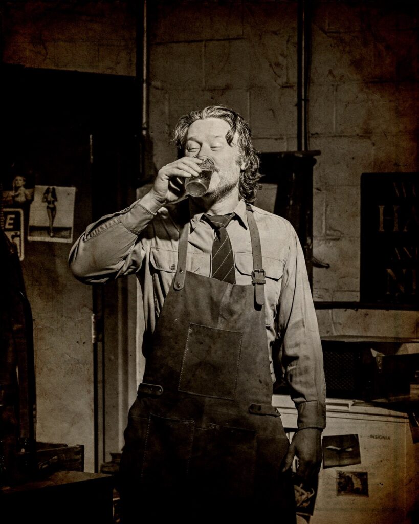 A man in an apron and shirt and tire drinks from a glass.