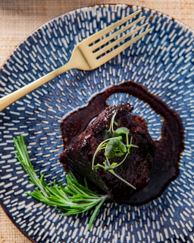 An overhead photo of a bite of meat garnished with micro greens and rosemary on a blue and white patterned plate with a gold fork.