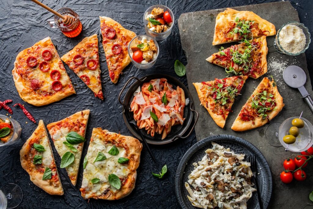 An overhead photo of different pizza slices and pasta dishes on slate and a textured black surface.