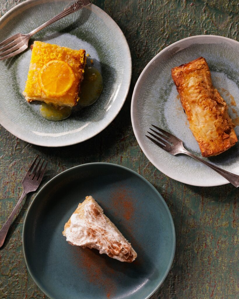 Three plates with desserts, one a slice of orange cake, one a pastry, and the other a slice of white cake with a cocoa dusting.