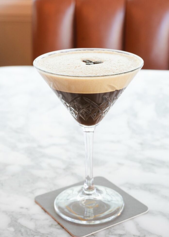 An espresso martini on a white marble table with a foam and three coffee beans for garnish.