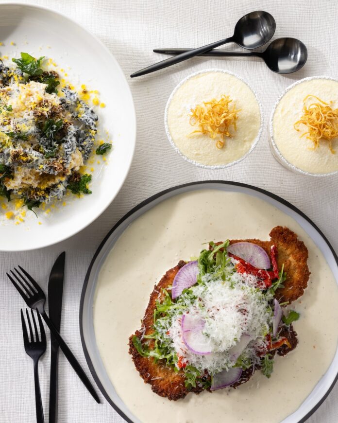 A white plate with a chicken parm stacked high with toppings beside a bowl of pasta and two dishes of lemon tiramisu.