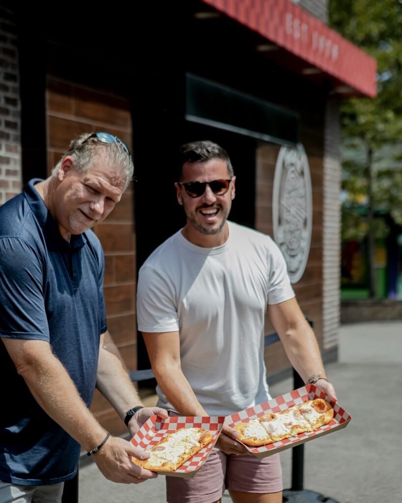 Two men hold out flatbread pizzas.